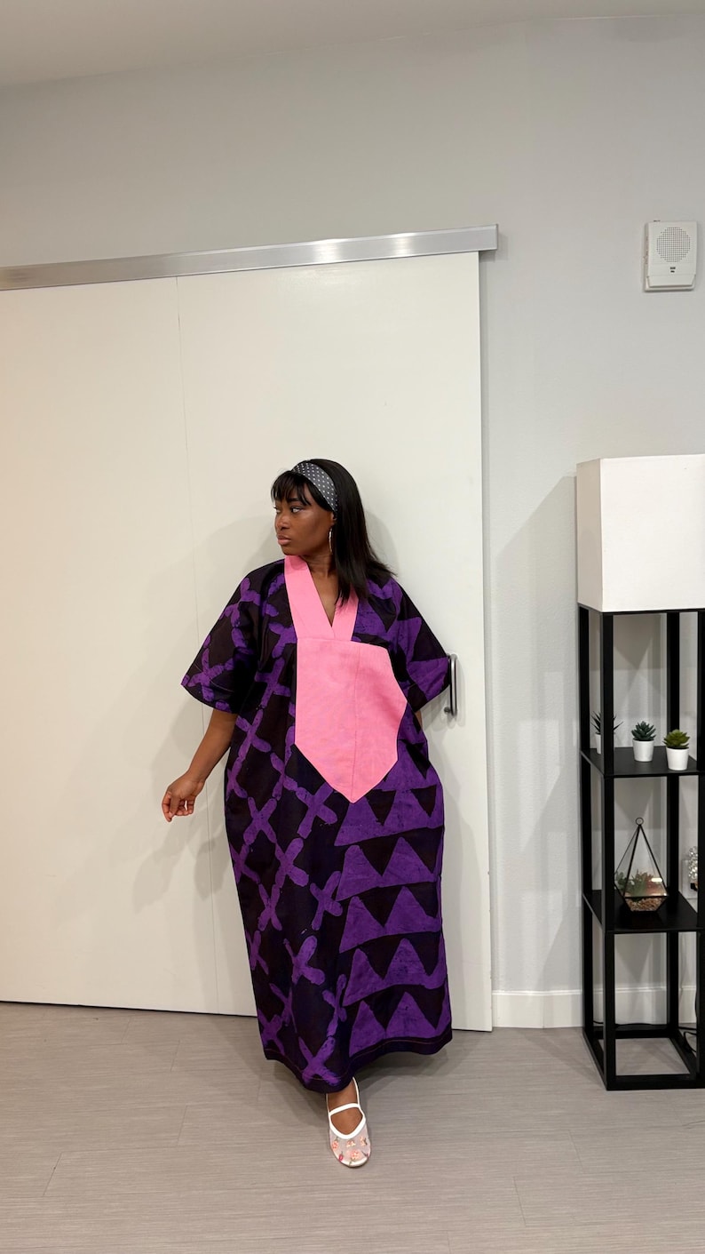 K&ouml;nnte beinhalten: Ein weit geschnittenes, bodenlanges Kaftankleid mit einem dunkelvioletten und schwarzen geometrischen Muster sowie einem markanten, einfarbigen rosa V-Ausschnitt-Einsatz. Das Kleid hat kurze, weite &Auml;rmel und eine lockere Passform, kombiniert mit wei&szlig;en Sandalen.