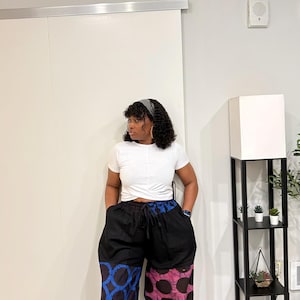May include: A white short-sleeved t-shirt with a front twist detail paired with wide-leg trousers. The trousers feature a solid black upper section with a drawstring waist and two distinct patterned lower legs: one with a blue circular motif and one with a pink and black circular pattern.