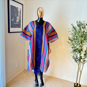 Pu&ograve; includere: Un abito caftano a righe colorato su un manichino. L'abito presenta uno scollo quadrato e un vivace motivo a righe rosa, blu, gialle e arancioni. L'abito ha un dettaglio di frange blu ed &egrave; esposto su un supporto nero.