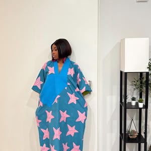 K&ouml;nnte beinhalten: Ein weit geschnittenes Kaftan-Kleid in Blaugr&uuml;n mit einem sich wiederholenden Muster aus hellrosa Sternen. Das Kleidungsst&uuml;ck hat einen einfarbigen blauen V-Ausschnitt und weite &Auml;rmel. Es reicht bis zu den Kn&ouml;cheln und wird mit wei&szlig;en Sandalen getragen.