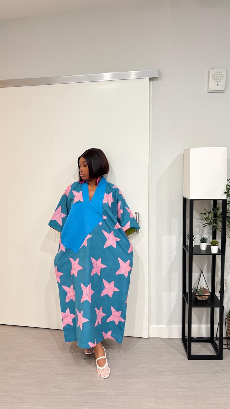 K&ouml;nnte beinhalten: Ein weit geschnittenes Kaftan-Kleid in Blaugr&uuml;n mit einem sich wiederholenden Muster aus hellrosa Sternen. Das Kleidungsst&uuml;ck hat einen einfarbigen blauen V-Ausschnitt und weite &Auml;rmel. Es reicht bis zu den Kn&ouml;cheln und wird mit wei&szlig;en Sandalen getragen.