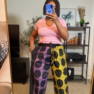 May include: Wide-leg pants with a unique design. One leg is black with large purple polka dots, and the other is yellow with black polka dots. The pants are paired with a light pink V-neck t-shirt.