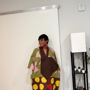 K&ouml;nnte beinhalten: Ein weit geschnittenes Midikleid mit einem gr&uuml;nen Oberteil mit rosa Dreiecksmustern und einem braunen Unterteil mit gelben und roten Kreismotiven. Das Kleidungsst&uuml;ck hat einen tiefen V-Ausschnitt mit einem braunen Einsatz.