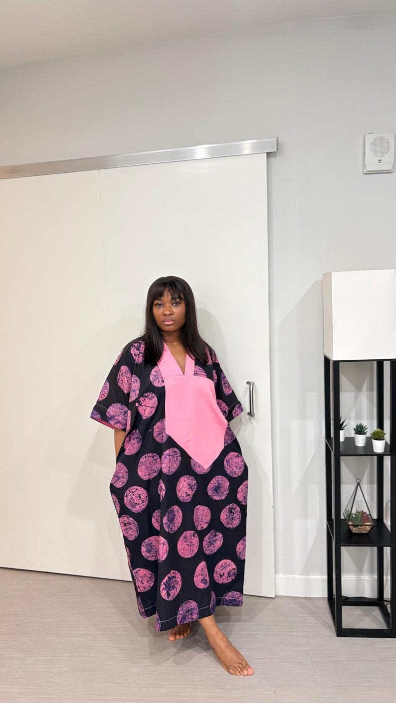 K&ouml;nnte beinhalten: Ein locker sitzendes schwarzes Kaftan-Kleid mit einem sich wiederholenden Muster aus texturierten rosa Kreisformen. Das Kleidungsst&uuml;ck verf&uuml;gt &uuml;ber einen einfarbigen rosa V-Ausschnitt, kurze weite &Auml;rmel und einen geraden Saum bis zur Wadenmitte.