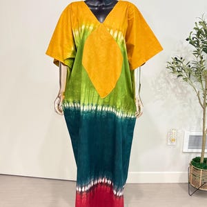 K&ouml;nnte beinhalten: Ein bodenlanges Kaftankleid mit V-Ausschnitt und kurzen &Auml;rmeln. Das Kleidungsst&uuml;ck zeigt ein Batikmuster mit horizontalen Streifen in Senfgelb, Limettengr&uuml;n, Dunkelwaldgr&uuml;n und einem roten Saum mit wei&szlig;en Akzenten.