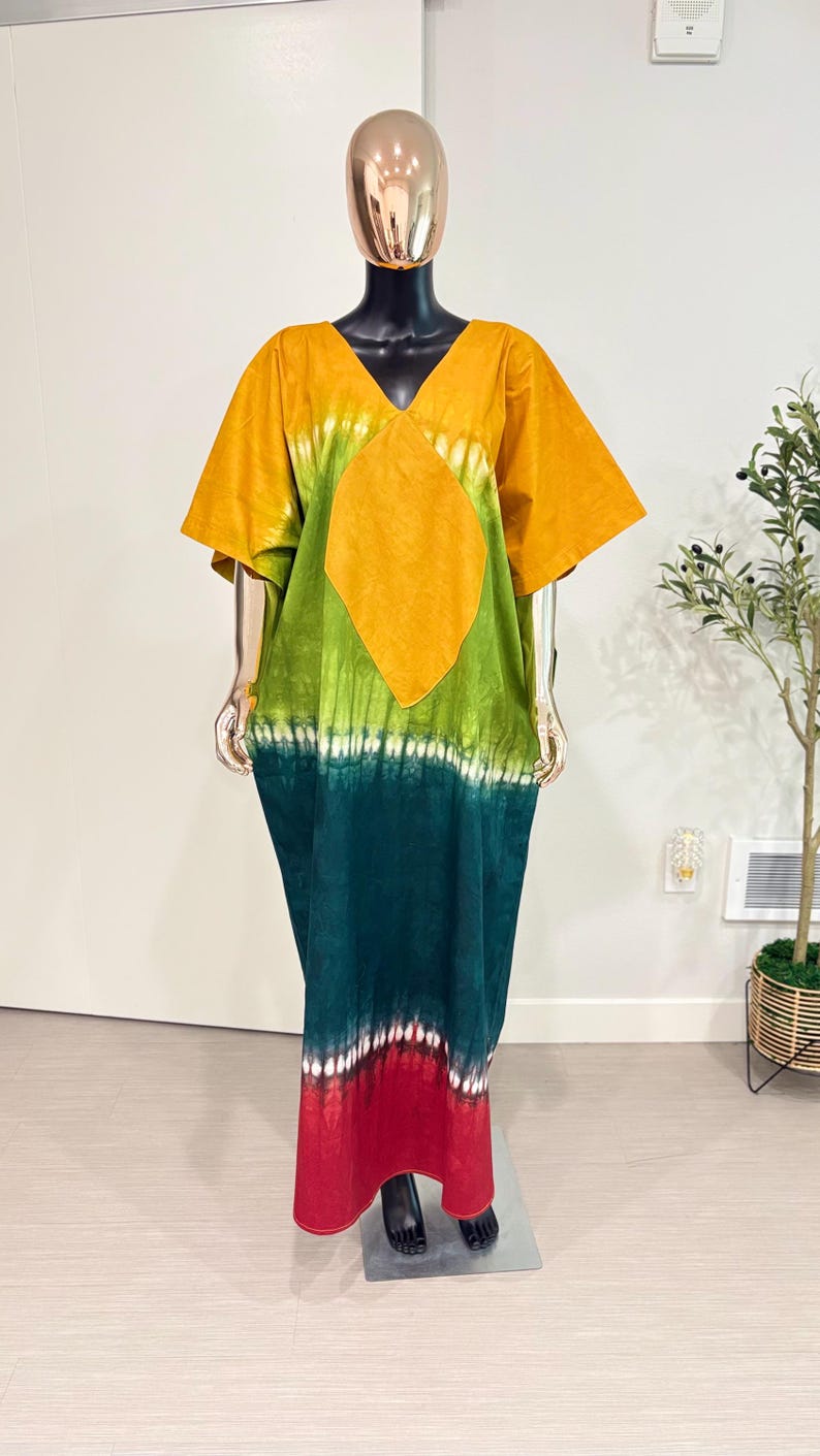 K&ouml;nnte beinhalten: Ein bodenlanges Kaftankleid mit V-Ausschnitt und kurzen &Auml;rmeln. Das Kleidungsst&uuml;ck zeigt ein Batikmuster mit horizontalen Streifen in Senfgelb, Limettengr&uuml;n, Dunkelwaldgr&uuml;n und einem roten Saum mit wei&szlig;en Akzenten.