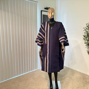 Pu&ograve; includere: Un abito blu navy in stile caftano con strisce verticali beige e blu. L'abito ha uno scollo a V, maniche larghe e una vestibilit&agrave; ampia. &Egrave; esposto su un manichino in una stanza ben illuminata.
