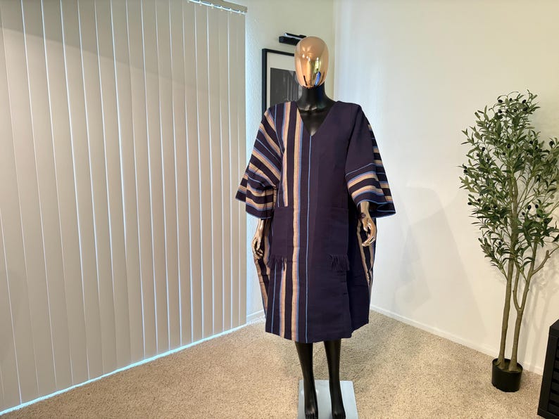 Pu&ograve; includere: Un abito blu navy in stile caftano con strisce verticali beige e blu. L'abito ha uno scollo a V, maniche larghe e una vestibilit&agrave; ampia. &Egrave; esposto su un manichino in una stanza ben illuminata.