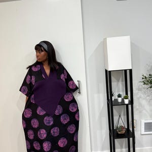 K&ouml;nnte beinhalten: Ein locker sitzendes, bodenlanges Kaftankleid mit schwarzer Grundfarbe und einem sich wiederholenden Muster aus gro&szlig;en, texturierten violetten Kreisen. Das Kleid hat einen einfarbigen, dunkelvioletten V-Ausschnitt und weite &Auml;rmel.