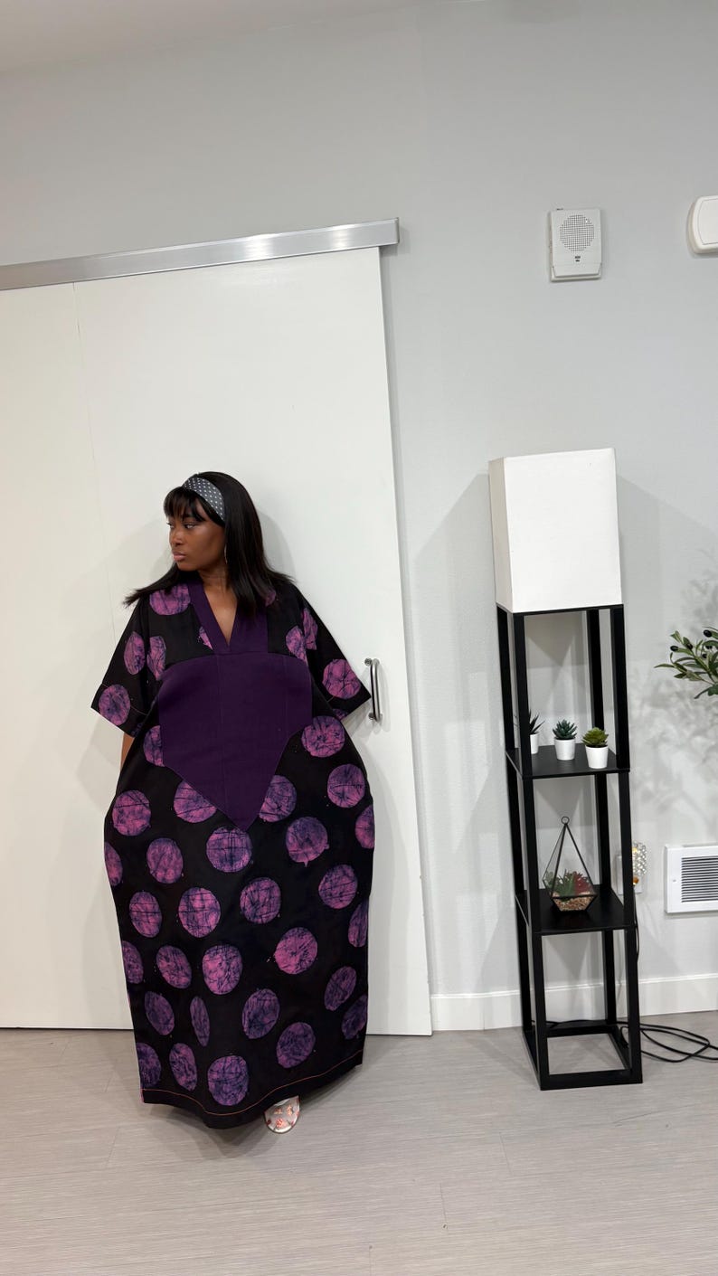 K&ouml;nnte beinhalten: Ein locker sitzendes, bodenlanges Kaftankleid mit schwarzer Grundfarbe und einem sich wiederholenden Muster aus gro&szlig;en, texturierten violetten Kreisen. Das Kleid hat einen einfarbigen, dunkelvioletten V-Ausschnitt und weite &Auml;rmel.