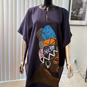 May include: Navy blue caftan dress with a keyhole neckline and short sleeves. The dress features a colorful painted design of a woman's face and head wrap. The artwork includes blue, orange, pink, and white accents.