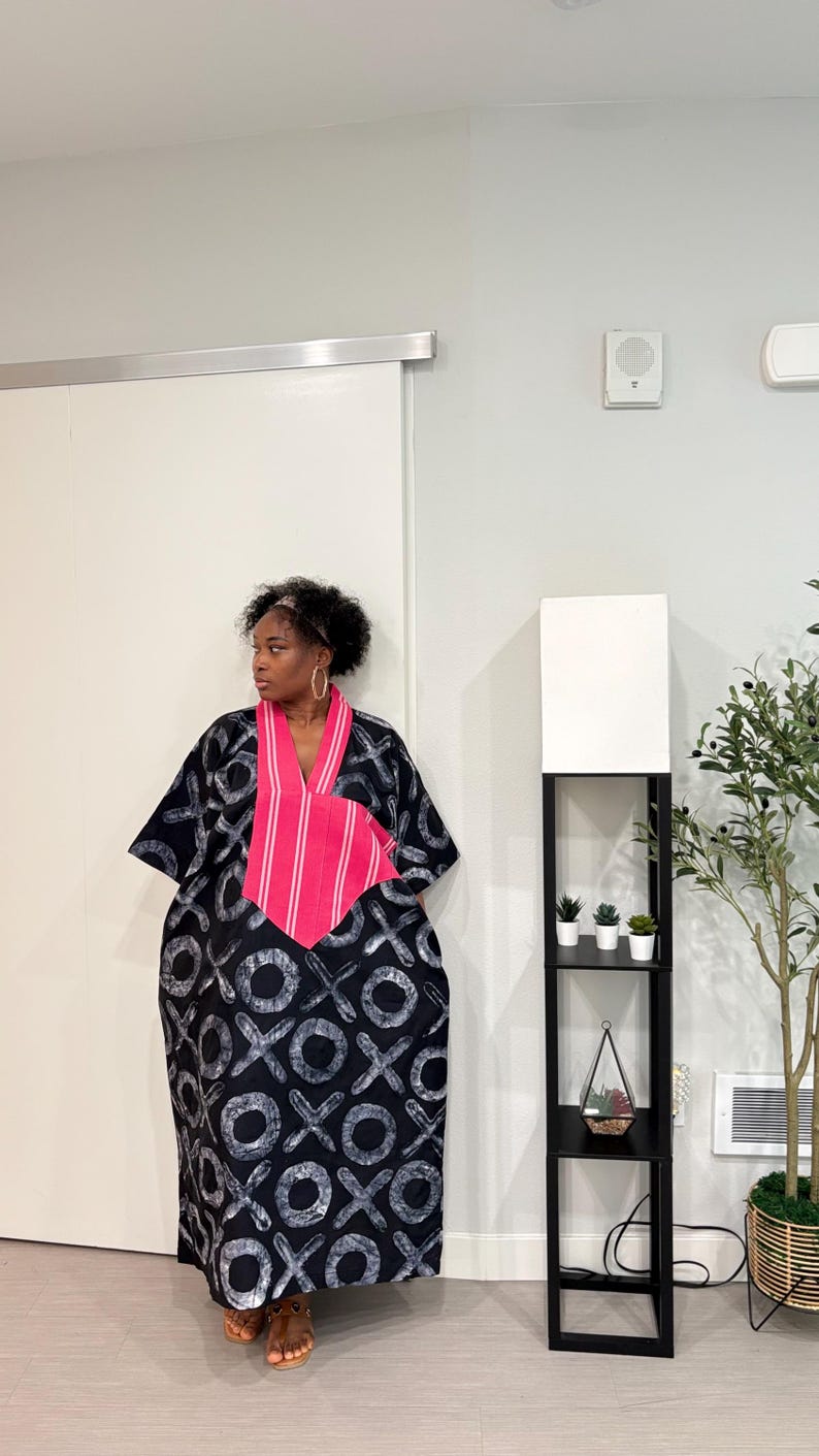 K&ouml;nnte beinhalten: Ein locker sitzender schwarzer Kaftan mit einem sich wiederholenden wei&szlig;en XOXO-Muster, akzentuiert durch einen leuchtend pinkfarbenen V-Ausschnitt mit vertikalen wei&szlig;en Streifen. Das Kleidungsst&uuml;ck hat weite kurze &Auml;rmel und einen bodenlangen Saum.