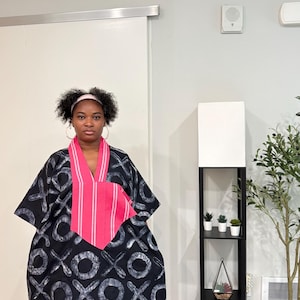 K&ouml;nnte beinhalten: Ein weit geschnittenes schwarzes Kaftan-Kleid mit einem sich wiederholenden wei&szlig;en X- und O-Muster. Das Kleidungsst&uuml;ck verf&uuml;gt &uuml;ber einen markanten V-Ausschnitt mit einem leuchtend pink-wei&szlig; gestreiften Stoffeinsatz auf der Brust.