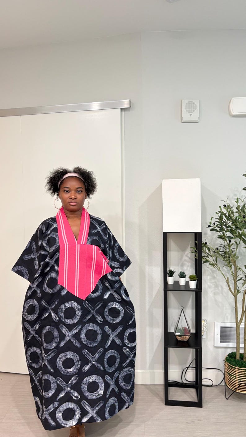 K&ouml;nnte beinhalten: Ein weit geschnittenes schwarzes Kaftan-Kleid mit einem sich wiederholenden wei&szlig;en X- und O-Muster. Das Kleidungsst&uuml;ck verf&uuml;gt &uuml;ber einen markanten V-Ausschnitt mit einem leuchtend pink-wei&szlig; gestreiften Stoffeinsatz auf der Brust.