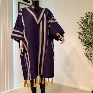 Pu&ograve; includere: Un abito tunica blu navy con strisce verticali beige, azzurre e oro. L'abito ha uno scollo a V e maniche corte, con nappe dorate lungo l'orlo. L'abito &egrave; esposto su un manichino.