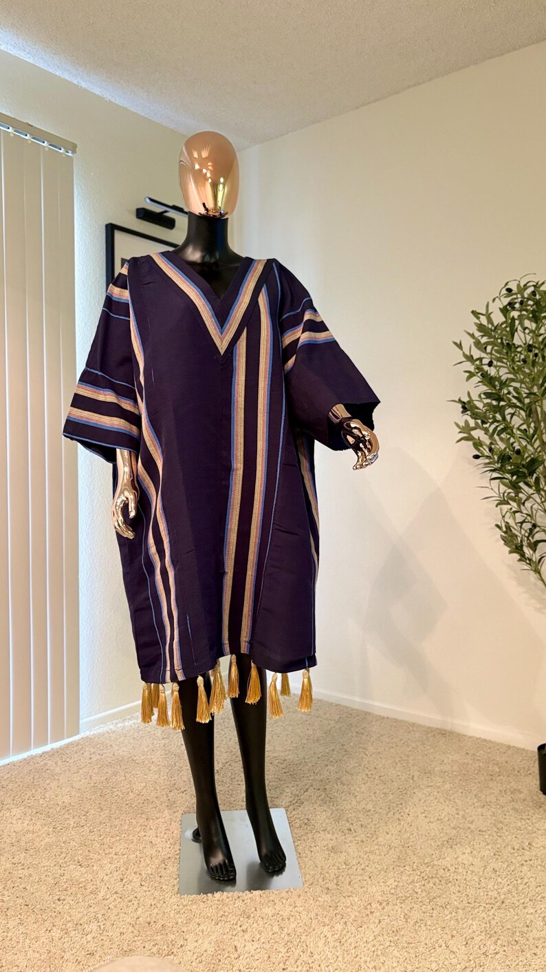 Pu&ograve; includere: Un abito tunica blu navy con strisce verticali beige, azzurre e oro. L'abito ha uno scollo a V e maniche corte, con nappe dorate lungo l'orlo. L'abito &egrave; esposto su un manichino.