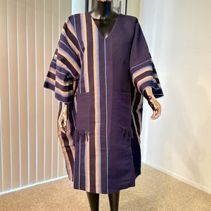 Pu&ograve; includere: Abito tunica blu navy con strisce verticali beige, blu e oro. L'abito presenta uno scollo a V, maniche corte e due tasche frontali con frange. L'indumento &egrave; esposto su un manichino.