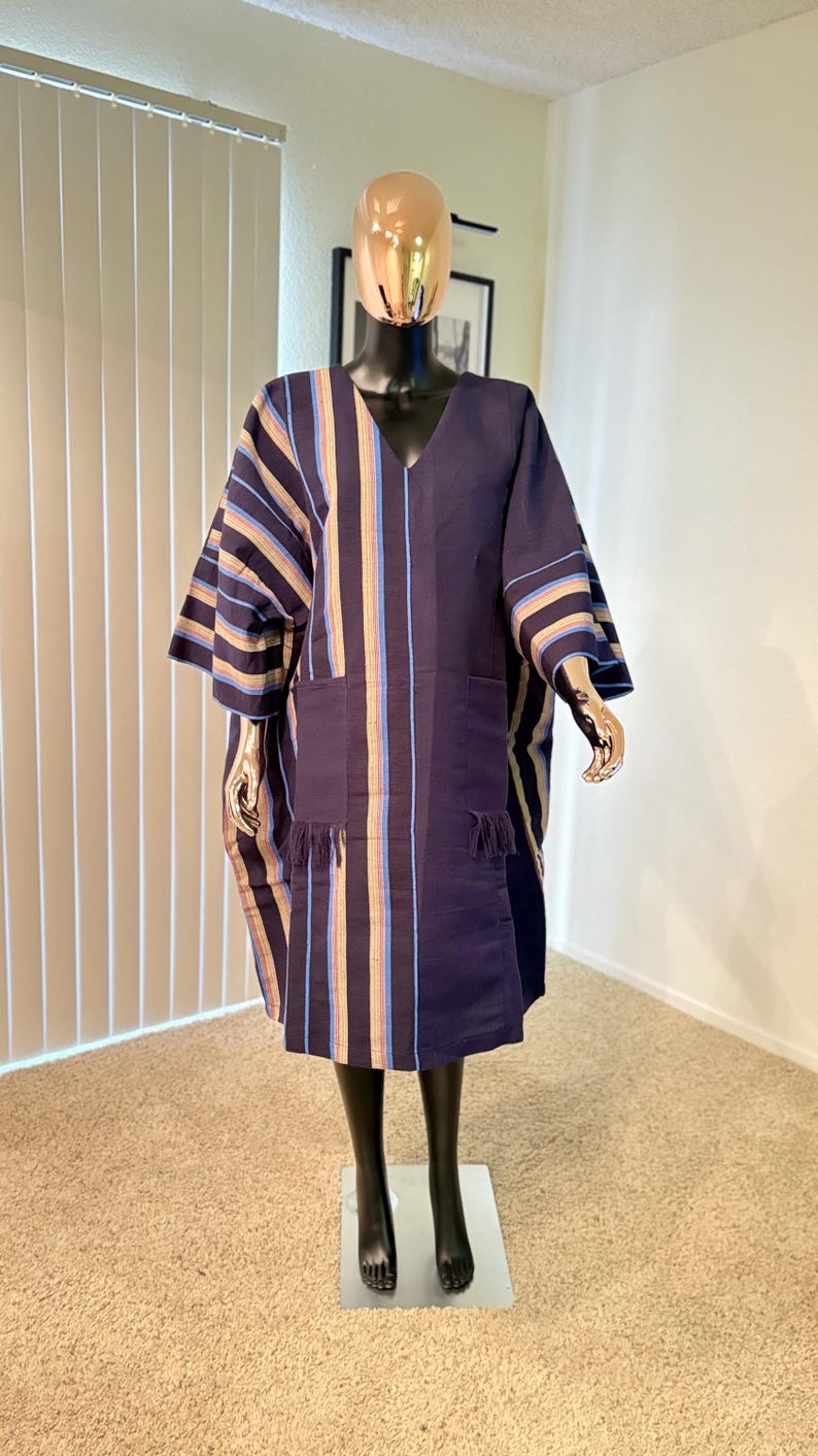 Pu&ograve; includere: Abito tunica blu navy con strisce verticali beige, blu e oro. L'abito presenta uno scollo a V, maniche corte e due tasche frontali con frange. L'indumento &egrave; esposto su un manichino.