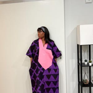 K&ouml;nnte beinhalten: Ein weit geschnittenes, kn&ouml;chellanges Kaftankleid mit tiefem V-Ausschnitt und einem einfarbigen rosa Einsatz. Das Kleidungsst&uuml;ck ist mit dunkelvioletten und schwarzen geometrischen Mustern, darunter wiederkehrende Dreiecke und Kreuze, sowie kurzen &Auml;rmeln versehen.