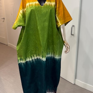 K&ouml;nnte beinhalten: Ein weit geschnittenes Kaftan-Kleid mit V-Ausschnitt und kurzen &Auml;rmeln. Der Stoff zeigt ein Batikmuster mit horizontalen Streifen in Senfgelb, Hellgr&uuml;n, Dunkelwaldgr&uuml;n und einem tiefroten Saum mit wei&szlig;en Akzenten.