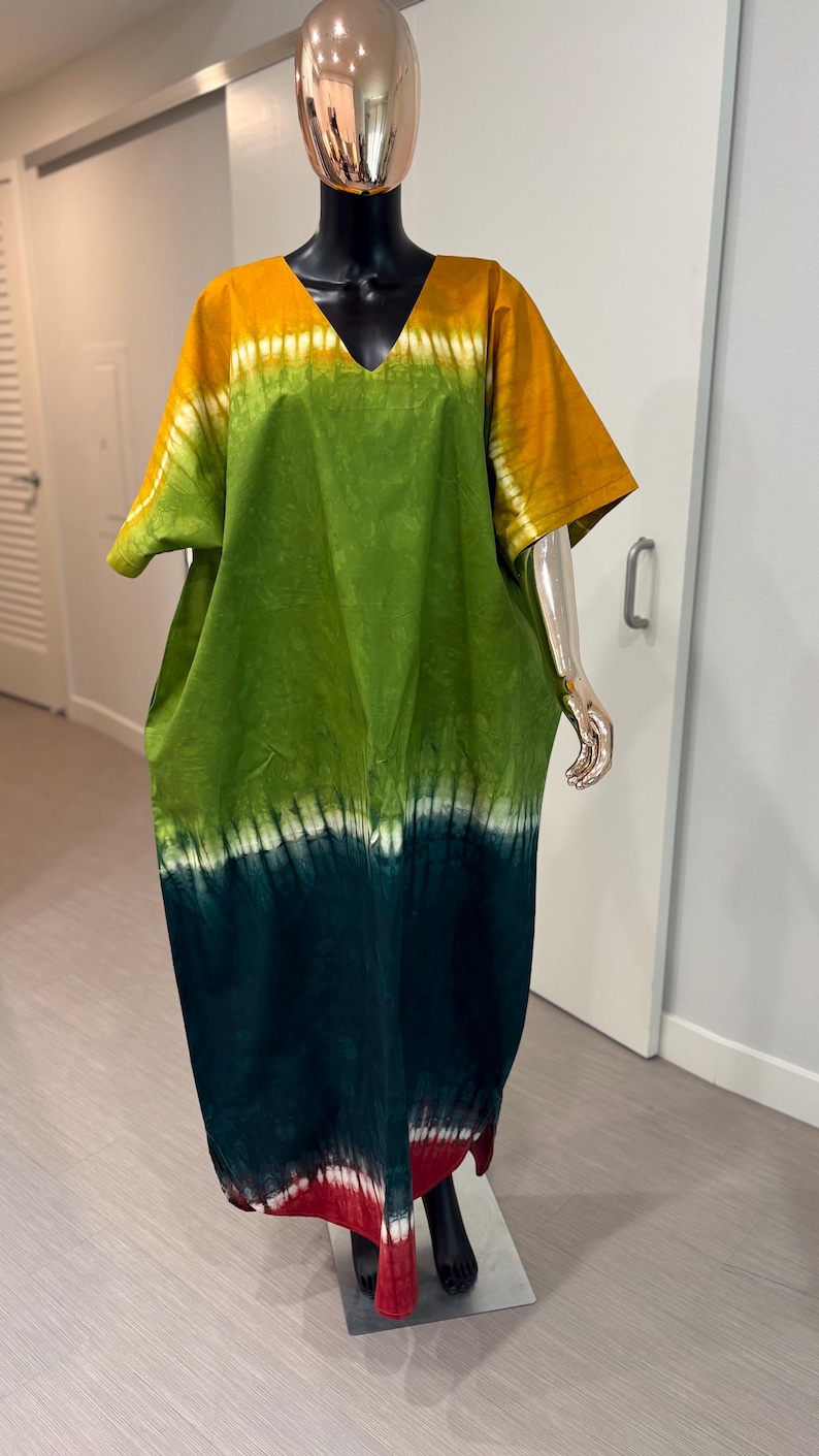 K&ouml;nnte beinhalten: Ein weit geschnittenes Kaftan-Kleid mit V-Ausschnitt und kurzen &Auml;rmeln. Der Stoff zeigt ein Batikmuster mit horizontalen Streifen in Senfgelb, Hellgr&uuml;n, Dunkelwaldgr&uuml;n und einem tiefroten Saum mit wei&szlig;en Akzenten.