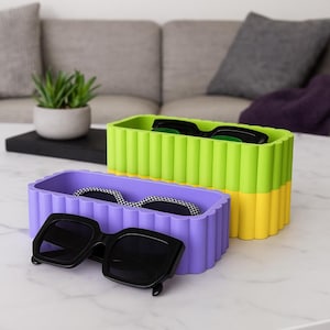 Può includere: Due contenitori rettangolari, uno lilla e uno verde lime, con un design a coste. Occhiali da sole neri sono appoggiati dentro e accanto ai contenitori. I contenitori sono su una superficie bianca.