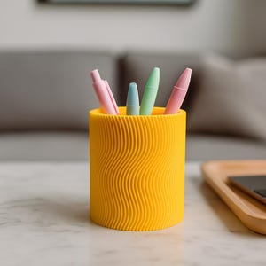 May include: A bright yellow cylindrical pen holder with a wavy, textured surface. The holder contains four pens in pastel colors: pink, blue, and green. The pen holder sits on a white marble surface.