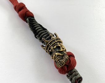 May include: A red, black, and gray lanyard with a brass skull pendant wearing a crown. The lanyard is made of braided cord and features a decorative knot. The skull pendant has intricate details.