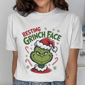 May include: White t-shirt with a Grinch graphic wearing a Santa hat, and the text "Resting Grinch Face." The design includes candy canes and snowflakes, ideal for the festive season.