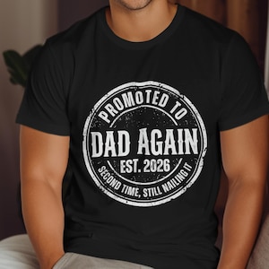 May include: Black t-shirt with a white circular graphic that reads "Promoted to Dad Again Est. 2026 Second Time, Still Nailing It". The shirt is short-sleeved and has a crew neck.
