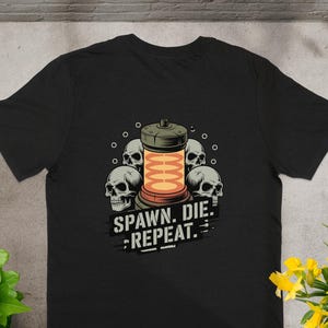 Puede incluir: Camiseta negra con un diseño gráfico de cuatro calaveras alrededor de un objeto cilíndrico brillante. El texto "SPAWN. DIE. REPEAT." está impreso debajo. La camiseta es de material suave.