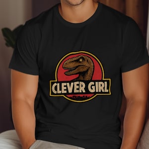 Può includere: T-shirt nero con grafica di dinosauro all'interno di un cerchio rosso e la scritta "CLEVER GIRL" in un riquadro bordato di giallo. Il dinosauro è marrone. La maglietta è a maniche corte.