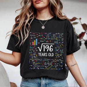 May include: Black t-shirt with a colorful math equation design. The shirt features the text "√196 YEARS OLD" in white, surrounded by mathematical formulas and symbols in various colors. A unique and educational apparel item.