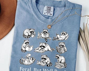 May include: A light blue t-shirt featuring a graphic of raccoons reading books in various poses. The text "Feral, But Well Read" is printed at the bottom. The shirt has a crew neck and short sleeves.