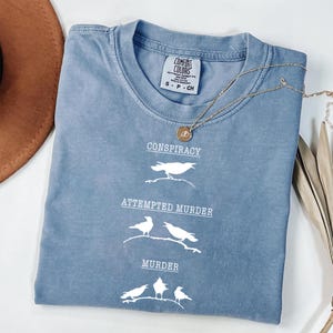May include: A blue Comfort Colors t-shirt with the words "Conspiracy", "Attempted Murder", and "Murder" in white, along with bird illustrations. A gold necklace with a pendant is draped across the shirt.