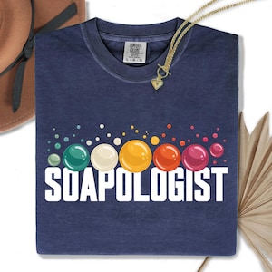 Puede incluir: Camiseta azul marino con la palabra "SOAPOLOGIST" en letras blancas. Encima del texto hay burbujas de colores en tonos verdes, amarillos, naranjas y rosas. Un collar dorado con un colgante de corazón está sobre la camiseta.
