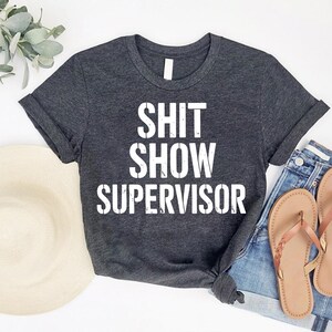 May include: Dark gray t-shirt with the words "SHIT SHOW SUPERVISOR" in bold white letters. The shirt is paired with denim shorts and brown sandals. A straw hat and greenery are also in the image.