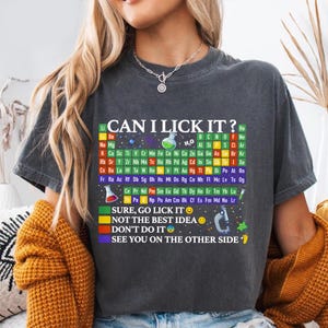 May include: Dark gray t-shirt with a periodic table design and the text "CAN I LICK IT?". Below the table are the phrases "SURE, GO LICK IT", "NOT THE BEST IDEA", "DON'T DO IT", and "SEE YOU ON THE OTHER SIDE".