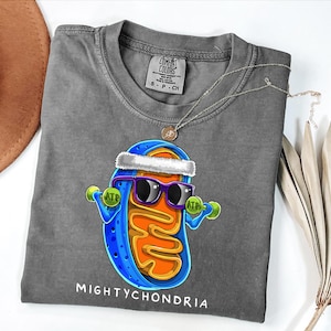 May include: Gray Comfort Colors t-shirt featuring a cartoon mitochondria character wearing sunglasses and a headband, holding dumbbells. The text "MIGHTYCHONDRIA" is printed below the graphic. A silver necklace is draped over the shirt.