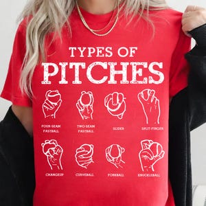 May include: Red t-shirt with the text "TYPES OF PITCHES" in white block letters. Below the text are illustrations of different baseball pitch grips, including four-seam fastball, slider, and knuckleball.