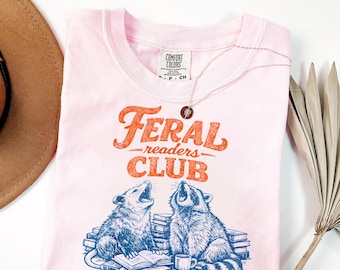 May include: A light pink t-shirt with the words "Feral Readers Club" in orange script. Below the text is a graphic of a raccoon and opossum, both looking up. A silver necklace with a small pendant is on the shirt.