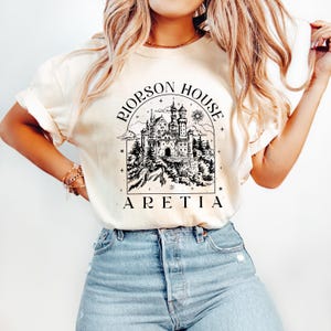 A woman wearing a white t-shirt with a graphic design of a castle and the text "Riope House Aretia" printed on it.