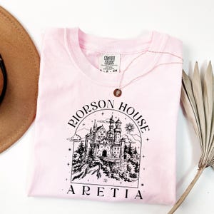 A pink t-shirt with a graphic design of a castle and the text "RIJORSON HOUSE ARETIA" printed on it, along with a necklace and a hat.
