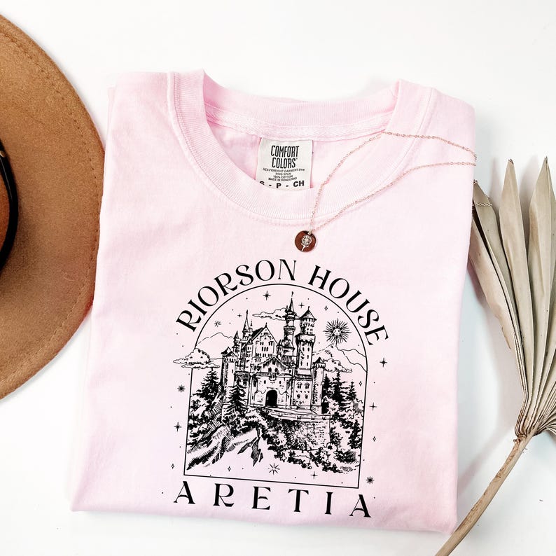 A pink t-shirt with a graphic design of a castle and the text "RIJORSON HOUSE ARETIA" printed on it, along with a necklace and a hat.