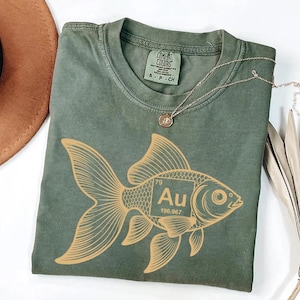 a green t-shirt with a gold fish graphic and the element Au (gold) printed on it, along with a necklace and a hat.