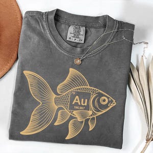 a gray t-shirt with a gold fish graphic and the text " Au" printed on it, along with a small gold pendant. The t-shirt is laid out on a white surface, and there is a brown hat and some dried leaves nearby.