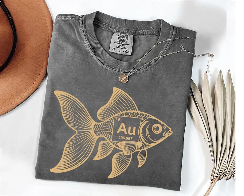 a gray t-shirt with a gold fish graphic and the text " Au" printed on it, along with a small gold pendant. The t-shirt is laid out on a white surface, and there is a brown hat and some dried leaves nearby.
