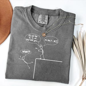 May include: Gray Comfort Colors t-shirt with a crew neck and the text "It's not the V=V₀at that kills you..." and "It's the F = MΔV". A gold necklace is draped on the shirt. The shirt has a small tag that says "Comfort Colors".