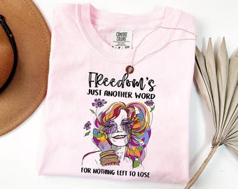May include: A light pink t-shirt with the text "Freedom's Just Another Word For Nothing Left To Lose" and a colorful graphic of a woman's face. A gold necklace is draped over the shirt.