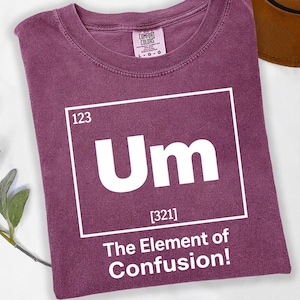 May include: A maroon t-shirt with a white graphic resembling a periodic table element. The graphic reads "Um" with the number "123" above and "[321]" below. The text "The Element of Confusion!" is printed below the graphic.